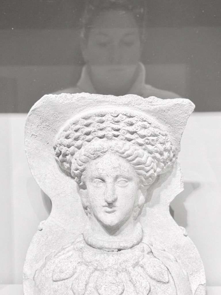 Self-portrait of Ashley reflected in glass behind an Etruscan sculpture